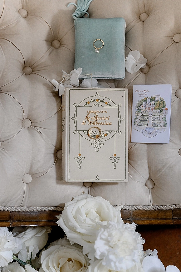 Wedding rings with engagement ring in a blue velvet ring box beside an illustrated invitation, white roses and petals on a chair