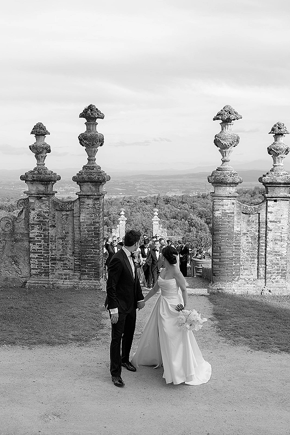 Couple portrait of bride and groom holding hands, bride looking back in strapless dress with bouquet by stone gate as guests cheer under cloudy sky