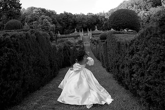 Bridal portrait of a bride running in gown, her long train billowing along a gravel path in a formal garden with hedges and stone stairs