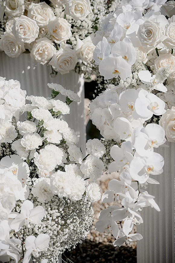 Wedding floral arrangement of white wedding flowers with roses, orchids, carnations, and baby’s breath on pedestal stands in sunlit greenery