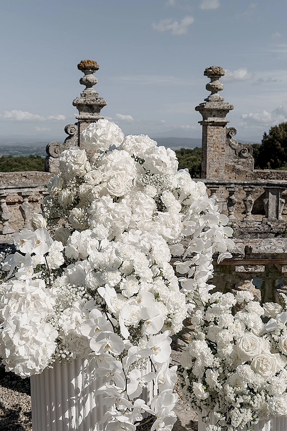 Wedding floral arrangement of white wedding flowers with roses, orchids, hydrangeas and greenery in a pedestal urn on a stone terrace overlooking hills