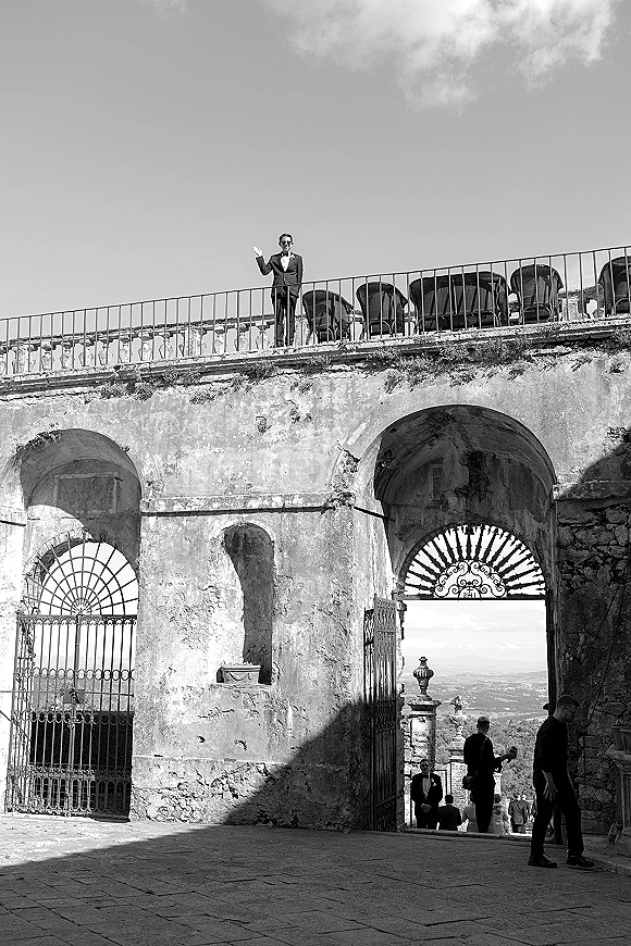 Groom portrait of a groom in tuxedo wearing sunglasses, leaning on a railing in a courtyard with stone arches and open doorway behind.