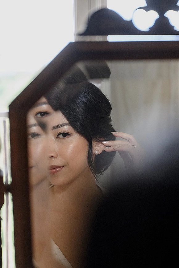 Bridal portrait of a bride mirror reflection as she puts on pearl stud earrings, engagement ring visible in soft window light indoors