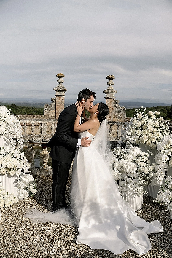 Wedding kiss portrait of bride and groom kissing, her cathedral veil and long train flowing on a stone terrace with mountain view
