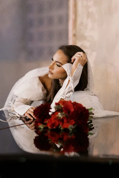 Bridal portrait of a bride with veil in a lace long-sleeve wedding dress, holding a deep red bouquet beside a stone wall in soft light