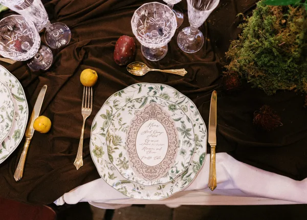 Reception tablescape wedding place setting with green floral china, calligraphy menu on plate, gold flatware, crystal glasses, fruit, and moss greenery