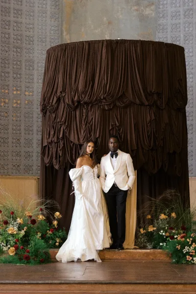 Couple portrait of bride in puff-sleeve corset gown and groom in white jacket with black bow tie before brown draped backdrop and roses