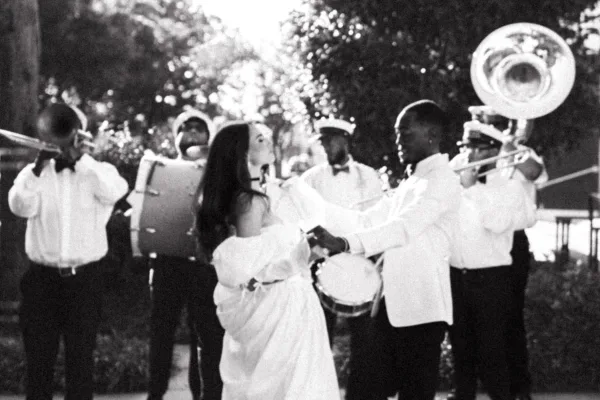 Wedding second line with bride leading a brass band parade, trombone and tuba players in uniforms, dancing outdoors among trees in daylight