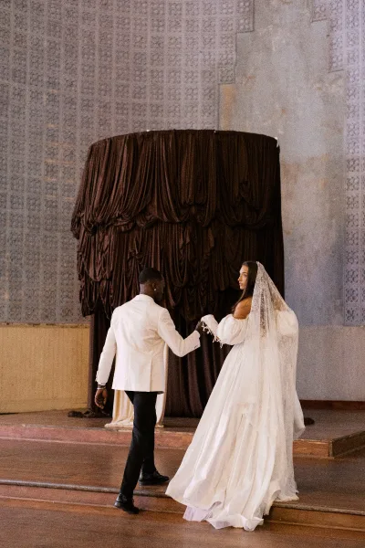 Couple portrait of bride and groom holding hands, bride glancing over her shoulder in veil and lace sleeves on indoor stage steps