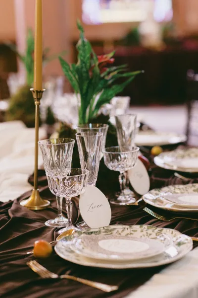 Reception tablescape wedding table setting with gold taper candle in brass holder, crystal glassware, patterned plates, and greenery on dark linen
