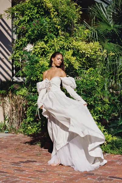 Bridal portrait of a bride in an off the shoulder wedding dress with puff sleeves, twirling on a sunlit brick walkway by palm greenery
