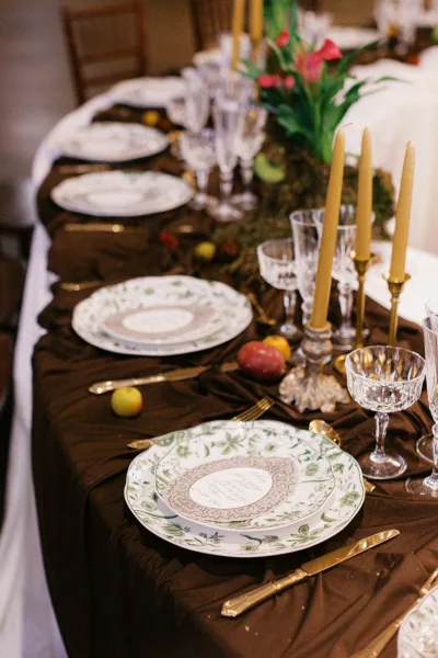 Reception tablescape with brown tablecloth, gold flatware, patterned plates, coupe and crystal glasses, taper candles, fruit, and greenery centerpiece indoors