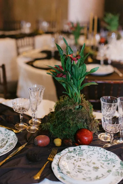 Reception tablescape with a wedding table centerpiece of moss, calla lilies, fruit, patterned plates, gold flatware, and crystal goblets indoors