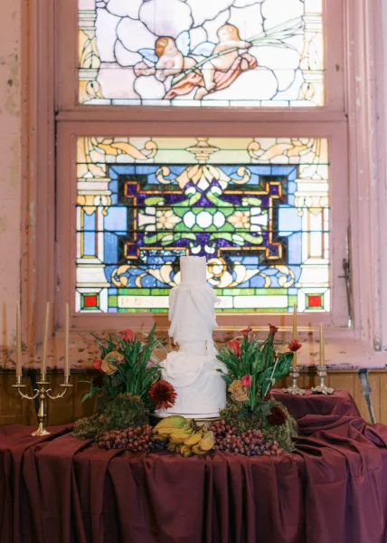 Wedding cake table with tiered wedding cake, gold candlesticks, calla lilies, and fruit display before a stained glass window backdrop