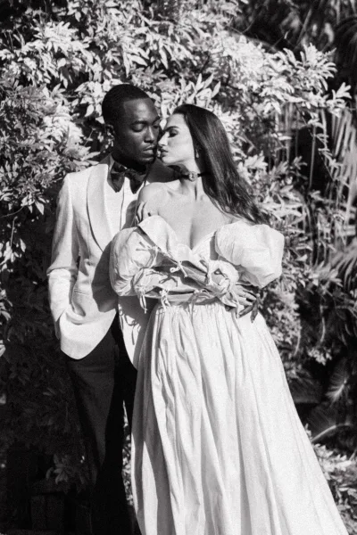 Couple portrait in a black and white wedding portrait, groom in white tuxedo embraces bride holding bouquet amid lush garden foliage