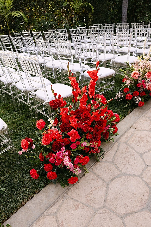 Ceremony aisle flowers in red roses and pink blooms line a stone garden walkway beside clear chiavari chairs on a grassy lawn