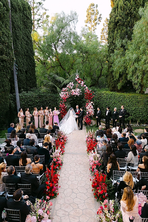Wedding ceremony with bride and groom at a floral arch, long veil and wedding party beside a flower-lined stone aisle in a garden