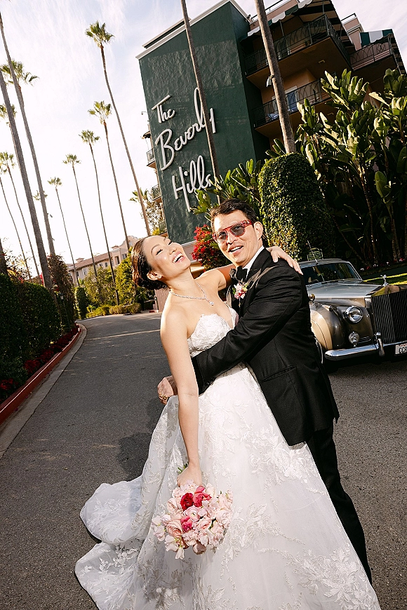 Couple portrait of bride and groom hug beside a vintage car, bride laughing in strapless lace dress as he wears sunglasses near palm-lined hotel driveway