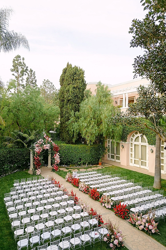 Outdoor ceremony setup with a garden wedding ceremony aisle lined with lush florals, leading to a floral arch by an ivy-covered building