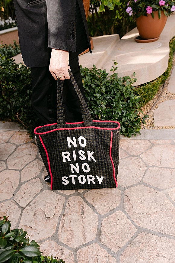 Wedding tote bag with custom text lettering held by a person in a black suit, wedding ring visible, on a stone patio by greenery