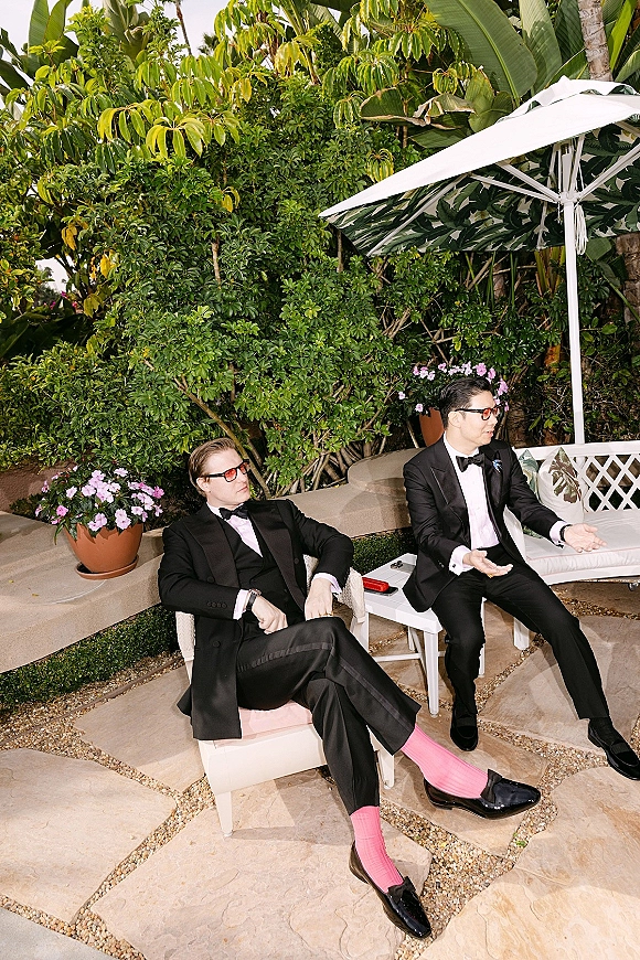 Groomsmen portrait of men in black tuxedos and sunglasses lounging in chairs, showing pink socks and loafers on a tropical patio