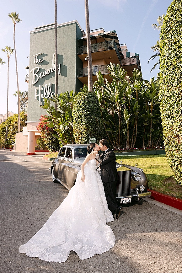 Wedding kiss portrait of bride and groom kissing beside a vintage luxury car, her strapless lace dress and train outside a palm-lined hotel exterior