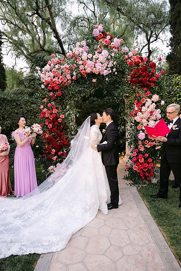Ceremony kiss as bride and groom embrace beneath a rose and greenery floral arch, her long veil trailing on a stone garden path