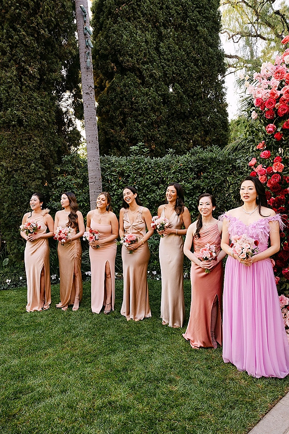 Bridesmaids group photo with bridesmaids holding bouquets in neutral satin dresses, standing on a garden lawn before a pink floral arch