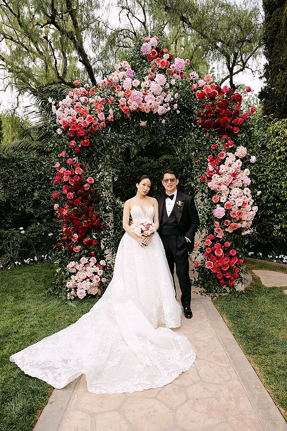 Couple portrait of bride in a strapless lace gown with bouquet beside groom in tuxedo and sunglasses under a floral arch in a garden