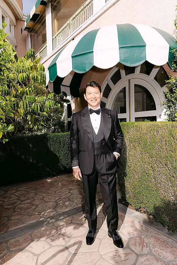 Groom portrait in a black tuxedo with bow tie and boutonniere, standing full-length by a striped awning and arched window on a stone walkway