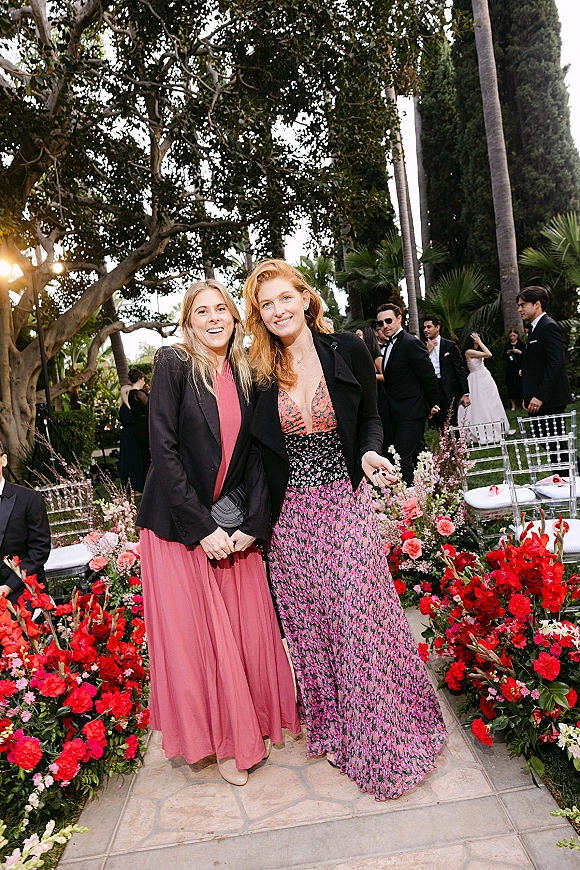 Wedding guest portrait in a floral midi dress with a black blazer and clutch, standing by clear chairs in a lush garden ceremony setting