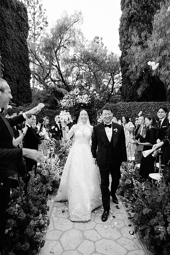 Recessional moment as bride and groom walk down the aisle, bouquet and veil flowing, guests cheering in an outdoor garden setting