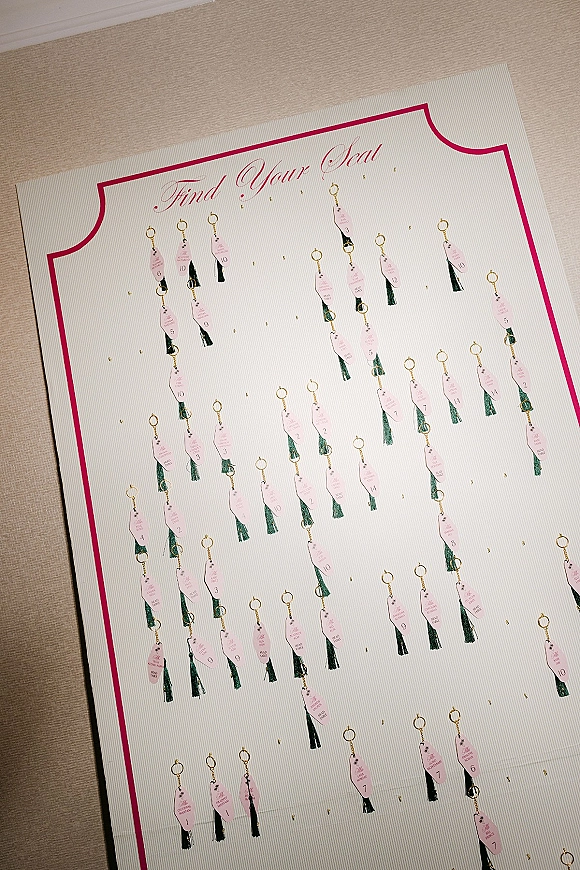 Escort card display with pink keychain tags and tassel escort cards on gold hooks beneath a printed header sign against a neutral wall
