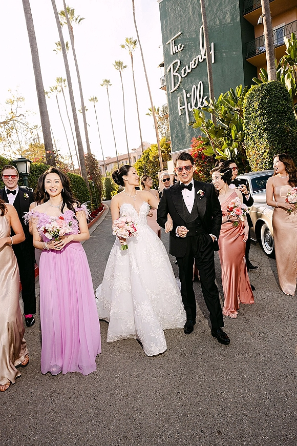 Wedding party portrait with bride and bridesmaids in pink dresses, holding bouquets and wearing sunglasses by a vintage car and palm trees