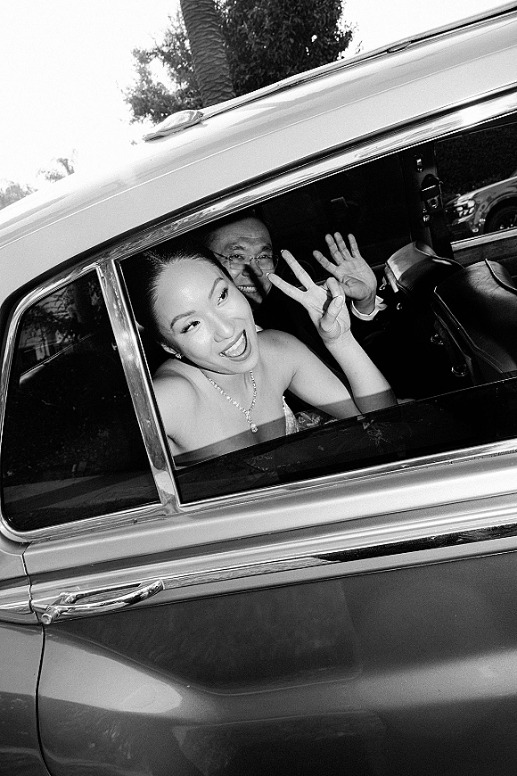 Wedding getaway car with bride leaning out the open window in a strapless dress, bridal necklace and earrings visible on a tree-lined street