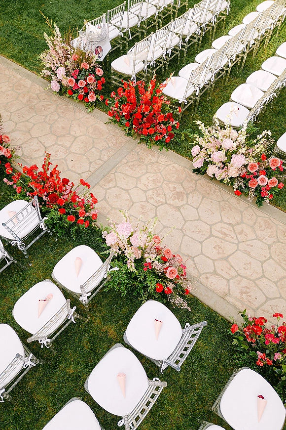 Ceremony aisle decor with outdoor ceremony aisle lined in red and pink florals, clear chiavari chairs, petal cones, and programs on a stone walkway
