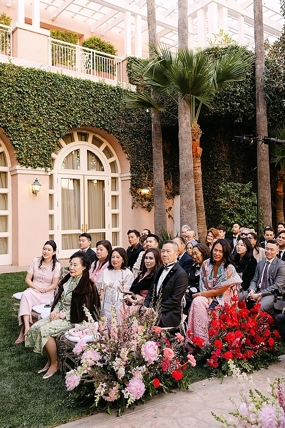 Wedding guests seated on clear acrylic chairs in formal attire, watching a courtyard ceremony with red and pink floral accents