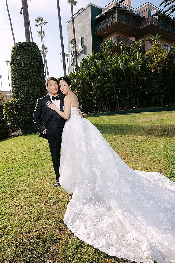 Couple portrait of bride hugging groom on a lawn, her strapless lace gown with long train beside his tuxedo amid palm trees and hotel balconies
