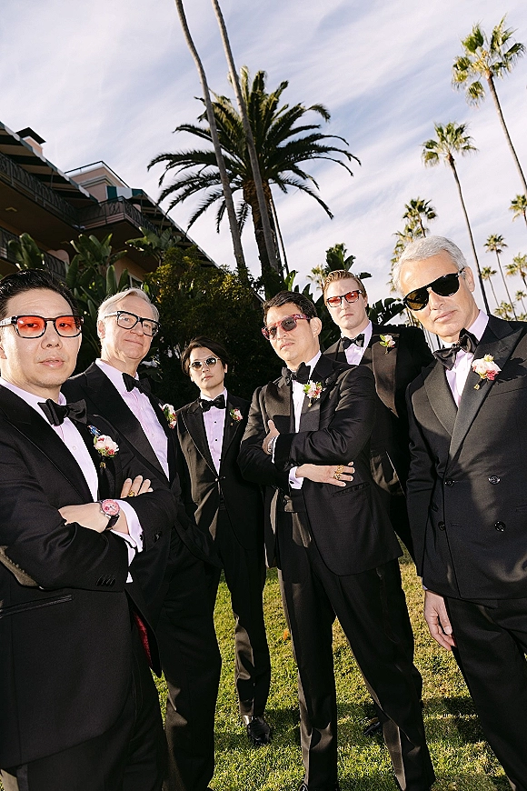 Groomsmen portrait of men in tuxedos wearing sunglasses, black tuxedos and boutonnieres on a lawn with palm trees and hotel backdrop