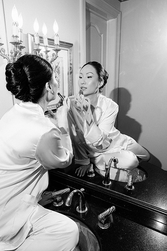 Bridal getting ready as the bride applying lipstick in a silk robe, seen in the bathroom mirror by a vanity with warm wall sconce light