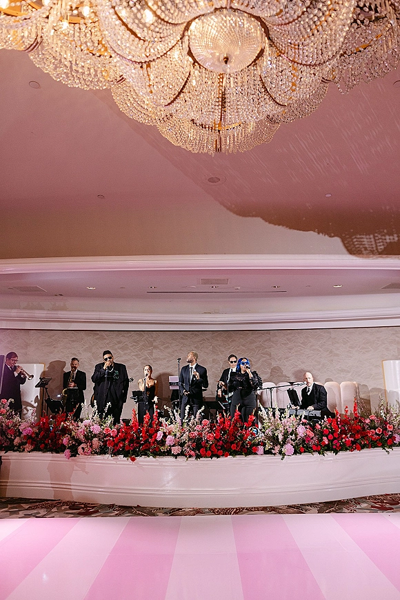 Wedding band performing with musicians and microphones on a stage riser with floral arrangement beneath a crystal chandelier in a ballroom