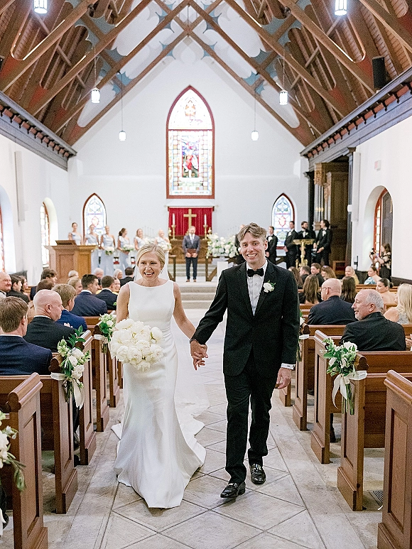 Wedding recessional as bride and groom walk hand in hand down a church aisle, bouquet and tuxedo amid ribbons, pew florals, stained glass windows