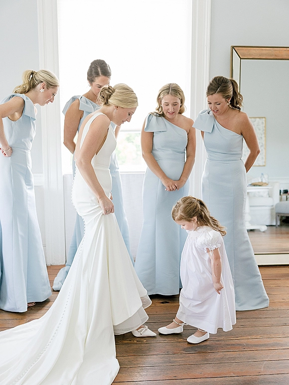 Bride with bridesmaids in blue dresses gathered by a bright window as she checks her long-train gown, with a flower girl in white nearby.