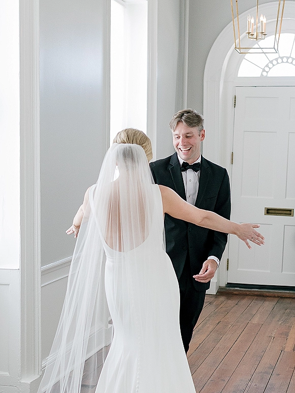 Wedding first look as bride in sleek dress and veil approaches groom in black tuxedo and bow tie in bright hallway with tall windows