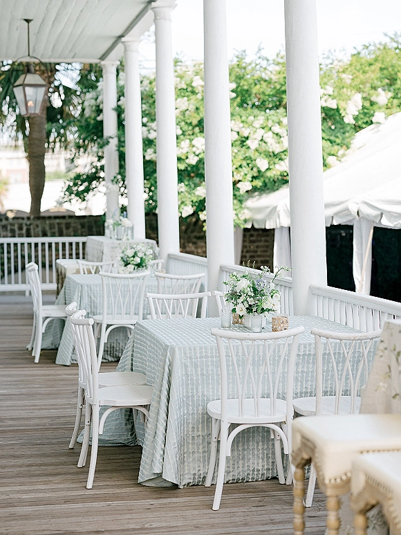 Reception tablescape with an outdoor reception table dressed in soft blue linens, white chairs, candles, and floral centerpieces on a covered porch