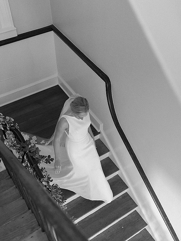 Bridal portrait of a bride walking down stairs in a minimalist wedding dress and veil, framed by indoor wooden staircase greenery garland.