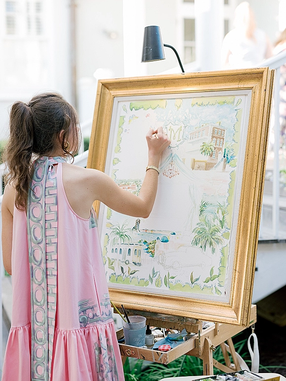 Live wedding painter at an easel creating wedding live painting on a gold-framed canvas beside a white building and greenery outdoors