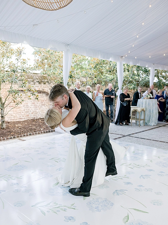 First dance kiss as bride and groom dip on the dance floor under string lights in a draped tent, guests by brick wall and greenery