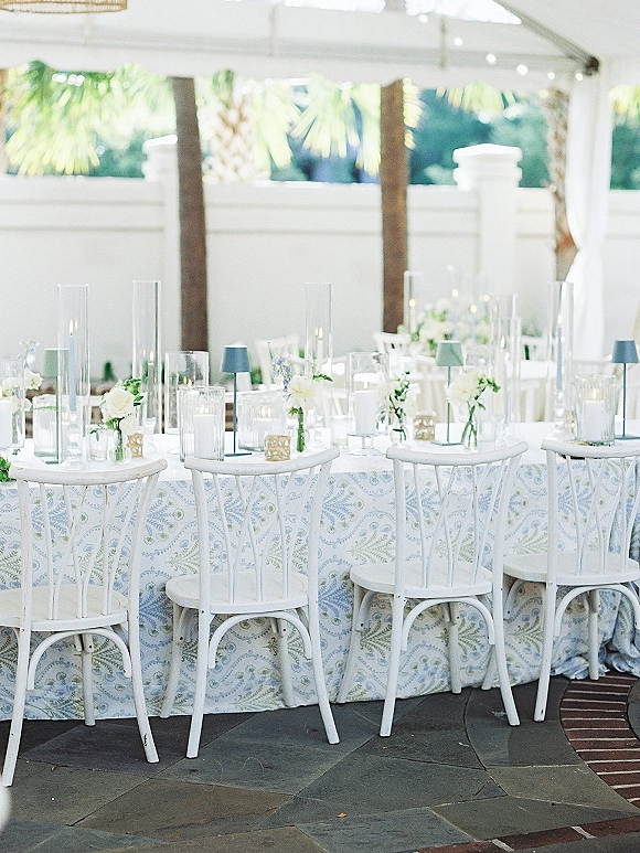Reception tablescape with blue and white patterned tablecloth, white roses and greenery bud vases, candles and glassware under a white tent canopy