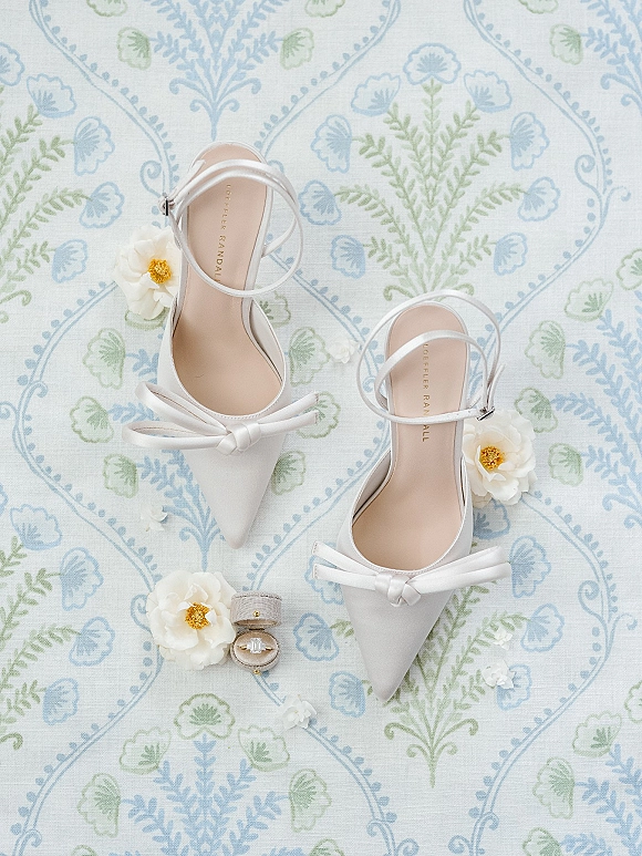 Bridal shoes flatlay featuring white satin bow heels with ankle straps, engagement ring and ring box on blue-green patterned fabric background